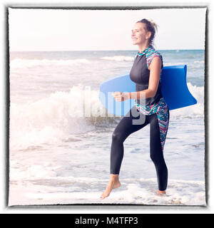 Donna bionda nella muta e scheda di nuoto di camminare sulla spiaggia Foto Stock