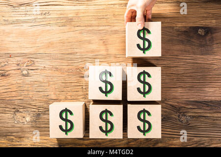 Close-up di mano di una persona di disporre un blocchetto in legno con il simbolo del dollaro sulla scrivania Foto Stock