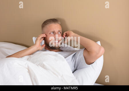 Turbato giovane uomo disteso sul letto che copre le sue orecchie per evitare rumore Foto Stock