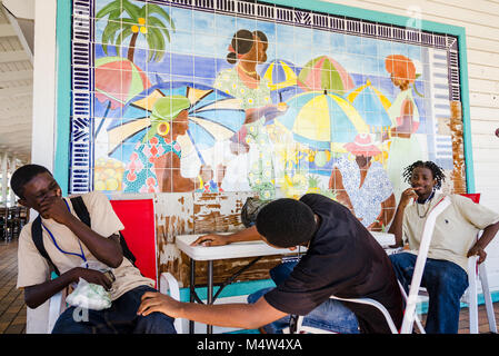 Marigot, San Martin. Tre giovani African American boys sedersi a un tavolo sotto un variopinto mosaico raffigurante un gruppo di donne in spiaggia. Foto Stock