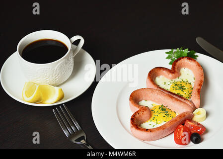 Uova strapazzate a forma di cuore su una piastra bianca con salsicce, pomodori verdi e il caffè con il limone. Foto Stock