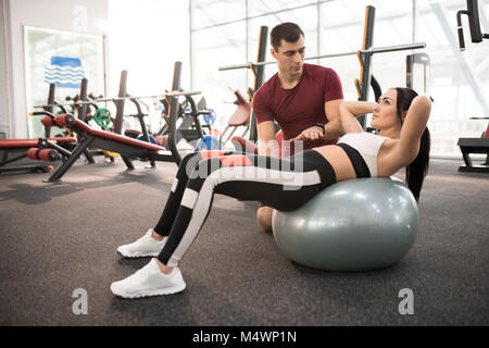 Vista laterale ritratto di allenatore muscolare aiutando giovane donna facendo esercizi sulla sfera di fitness in una palestra moderna, spazio di copia Foto Stock