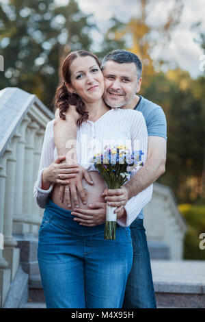 Giovane uomo abbracciando la sua moglie incinta. Egli detiene un bouquet di fiori nelle sue mani Foto Stock