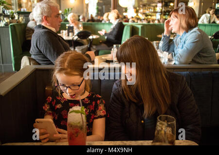 Madre e figlia di 8 anni in un affollato bar alla ricerca e riproduzione su Apple iPhone Foto Stock