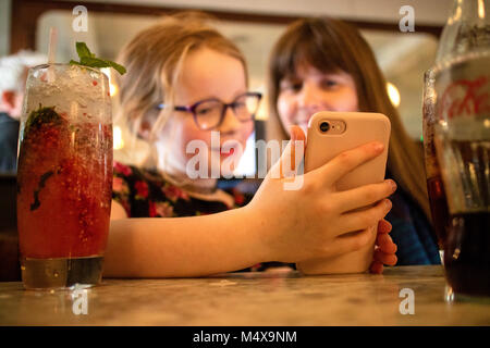 Madre e figlia di 8 anni in un affollato bar alla ricerca e riproduzione su Apple iPhone Foto Stock