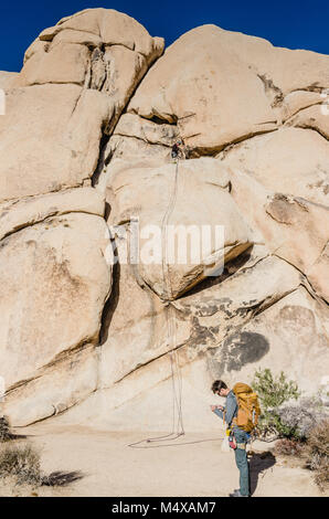 Giovane uomo cuscinetto pacco completo programma la sua arrampicata nella parte anteriore della corda ancorata sulla roccia di intersezione, la prominente 150-piede alto monolito monzonite. Foto Stock