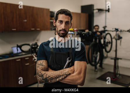 Lavoratore in piedi con le braccia incrociate in possesso di una chiave inglese in officina. Meccanica assemblaggio di biciclette in officina di riparazione. Foto Stock