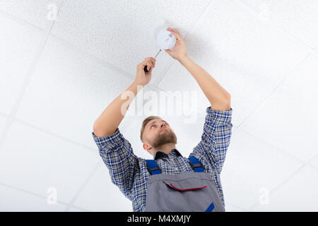 Basso Angolo di visione di un elettricista maschio fissaggio rivelatore di fumo mediante il cacciavite sulla parete di soffitto Foto Stock