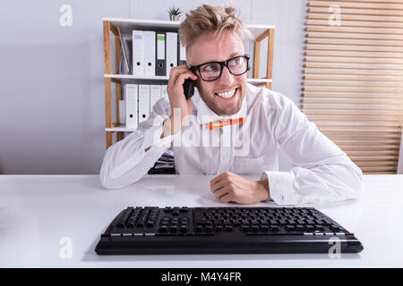 Felice giovane imprenditore parlando su Mobilephone con la tastiera sulla scrivania Foto Stock