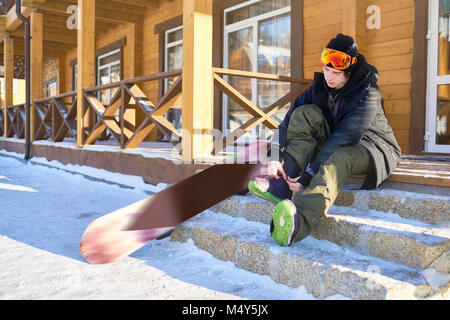 A piena lunghezza Ritratto di giovane moderno snowboarder mettendo su scarponi seduti sui gradini di chalet in legno a ski resort, spazio di copia Foto Stock