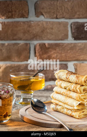 Pizzo sottili frittelle in pila i dolci fatti in casa sulla tavola di legno close-up con miele e tè di fiori dai boccioli secca di una rosa rossa. Cibo Concetto di vacanza Shrov Foto Stock