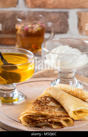 Pizzo sottili frittelle in pila i dolci fatti in casa sulla tavola di legno close-up con miele e panna acida e il tè di fiori dai boccioli secca di una rosa rossa. Concep alimentare Foto Stock