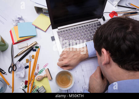 Close-up di esaurito imprenditore appoggiato il suo capo sul laptop tramite il disordine Desk al luogo di lavoro Foto Stock