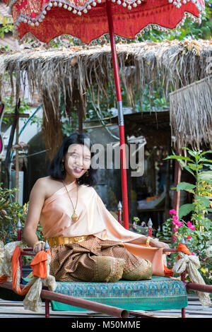 Giovane donna in tradizionali abiti tailandesi si siede sulla storica figliata. Ragazza Thai in costume storico rilassato sotto l'ombrello. Foto Stock