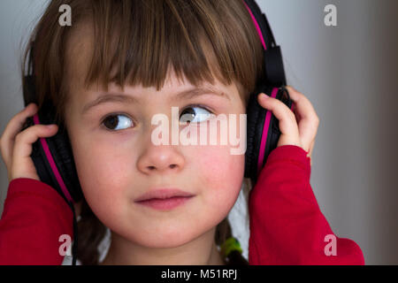 Foto di felice bambina carina bambina con grandi cuffie a casa. Gioiosa ragazza bambino ascoltare musica su grigio sfondo sfocato. Home, technolo Foto Stock