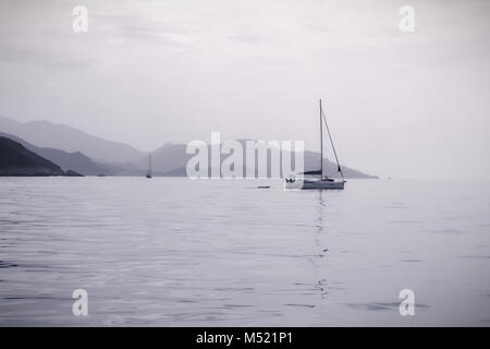 Barca a vela nei mari calmi con le montagne in distanza in monotono grigio scuro a whiteout. Le montagne della Grecia nel mar Mediterraneo off Corfù Foto Stock