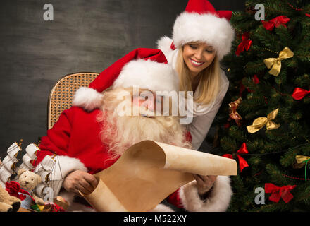 Ritratto di felice Babbo Natale seduto alla sua stanza di casa con la sua donna helper nei pressi di albero di Natale e grande sacco e la lettura della lettera di Natale o desiderate Foto Stock