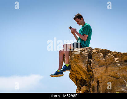 Teen ragazzo seduto su una scogliera e smartphone di lettura a Torrey Pines State riserva naturale vicino a San Diego, CA. Foto Stock