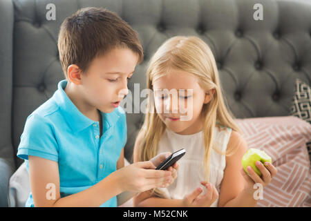 Piccolo fratello e sorella sul divano giocando al telefono cellulare. Ritratto di famiglia Foto Stock