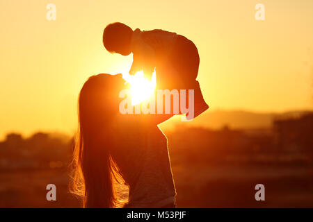 Retroilluminazione silhouette di una madre alzando la kid figlio al tramonto con il sole in medio Foto Stock