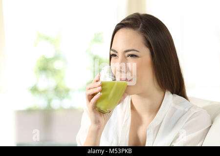 Donna felice di bere un verde un succo vegetale seduta su un divano nel salotto di casa Foto Stock