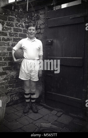 1920s, foto storiche, mostra un orgoglioso giovane ragazzo in piedi nel suo ingranaggio di calcio e stivali tenendo un calcio in pelle nel suo cortile, Inghilterra, Regno Unito. Foto Stock