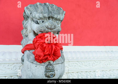 Tradizionale statua di pietra di un tutore cinese lion, indossando un grande arco rosso attorno al collo per festeggiare il nuovo anno cinese, pubblicato nella parte anteriore di un rosso Foto Stock