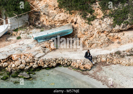 Una calma in attesa donna seduta su una roccia accanto a una barca da pesca stranged sulla spiaggia di Cala Pi sull'isola di Mallorca, Spagna. Foto Stock