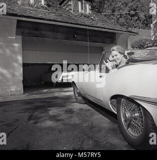 Anni sessanta, STATI UNITI D'AMERICA, storico, bella signora al volante del suo open-top Jaguar auto sportiva utilizzando i più recenti accessori per automobile, un dispositivo di controllo remoto che si apre automaticamente le porte di garage, Los Angeles, California. Foto Stock