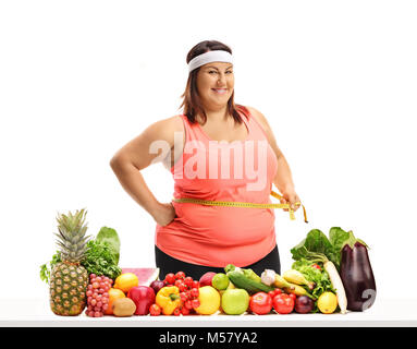 Donna sovrappeso tenendo un nastro di misurazione circa la sua vita dietro un tavolo con frutta e verdura isolati su sfondo bianco Foto Stock