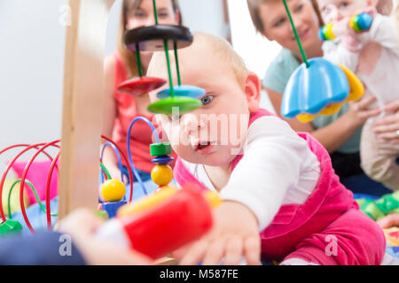 Carino bambina che mostra il progresso e curiosità cercando di raggiungere multicolore di giocattoli di legno, mentre è seduto sul pavimento con la madre e una famiglia ven Foto Stock