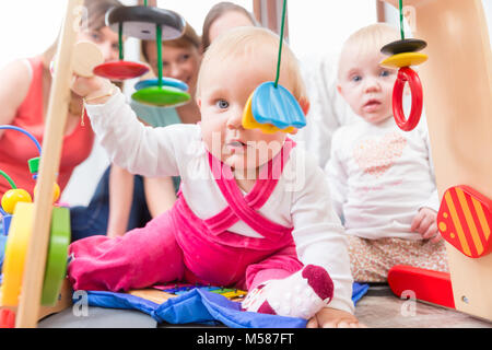 Carino bambina che mostra il progresso e curiosità cercando di raggiungere multicolore di giocattoli di legno, mentre è seduto sul pavimento con la madre e una famiglia ven Foto Stock