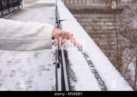 Primo piano della mano womans disegno di una linea in neve fresca sulla ringhiera in città il Parco in inverno Foto Stock