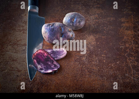 Fette di patate vitelotte, tre sezioni sul coltello e due patate intere su terreni accidentati sfondo scuro Foto Stock