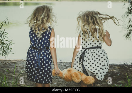 Due bambine con lunghi capelli biondi tenendo un orsacchiotto di peluche e giocattoli in esecuzione per l'acqua Foto Stock