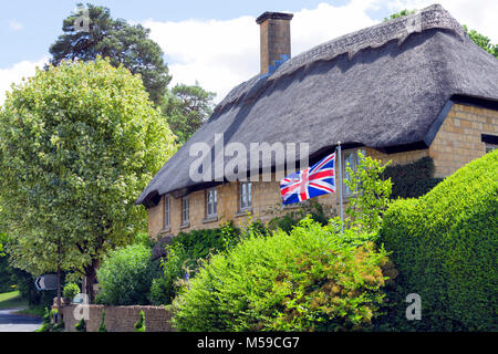 Il vecchio tetto di paglia golden cottage in pietra da un villaggio rurale road, con sventola bandiera britannica nel giardino anteriore . Foto Stock