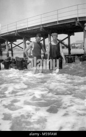 Una giovane coppia sta nel surf dalla Boardwalk mentre nuotare nell'Oceano Atlantico, ca. 1920. Foto Stock