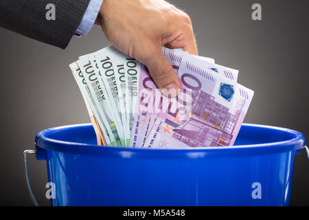 Ritagliate la mano dell'uomo d'affari gettando le banconote in euro in blu la benna contro uno sfondo grigio Foto Stock