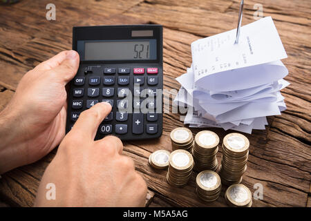 Immagine ritagliata di imprenditore utilizzando la calcolatrice con le monete e le fatture su un tavolo di legno Foto Stock