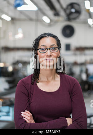 Donna con capelli castani con gli occhiali in piedi in officina di metallo, sorridente alla fotocamera. Foto Stock