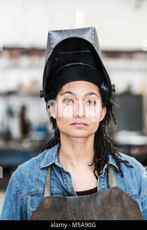 Ritratto di donna che indossa il grembiule e la maschera di saldatura permanente di metallo in officina, guardando la fotocamera. Foto Stock