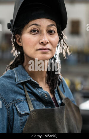 Ritratto di donna che indossa il grembiule e la maschera di saldatura in piedi in officina di metallo, sorridente alla fotocamera. Foto Stock