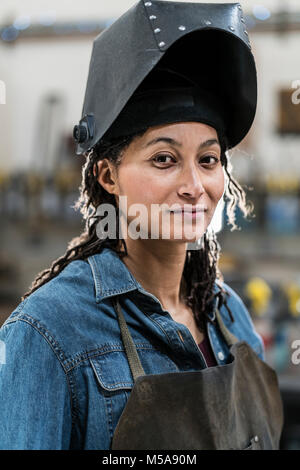 Ritratto di donna che indossa il grembiule e la maschera di saldatura in piedi in officina di metallo, sorridente alla fotocamera. Foto Stock