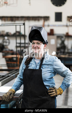 Ritratto di donna che indossa il grembiule e la maschera di saldatura in piedi in officina di metallo, sorridente alla fotocamera. Foto Stock