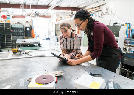 Due donne in piedi al banco da lavoro in metallo workshop, guardando a tavoletta digitale. Foto Stock