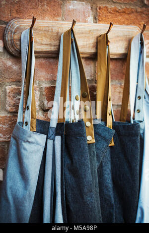 Denim grembiuli di lavoro appeso su una fila di ganci su un muro di mattoni. Foto Stock