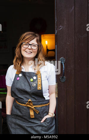 Donna con capelli lunghi marrone che indossa il grembiule e bicchieri in piedi in una porta aperta, sorridente. Foto Stock