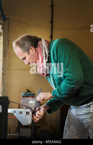 Un artigiano in un coltellinai workshop, tenendo un coltello da cucina e sagomatura e finitura del manico in legno con una smerigliatrice di superficie di una ruota rotante. Foto Stock