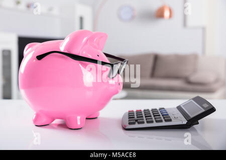 Close-up di Rosa Piggybank indossa occhiali con la calcolatrice sulla scrivania Foto Stock