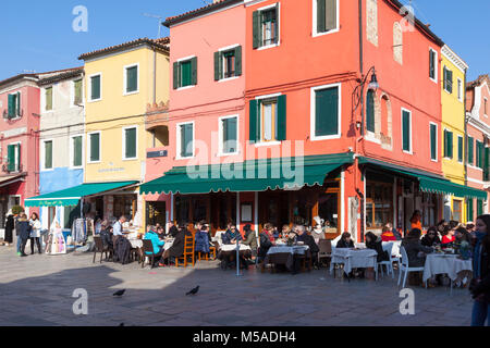 Isola di Burano, Venezia, Veneto, Italia. Persone a cena in un ristorante ad angolo su Via Baldassare Galuppi su una soleggiata giornata invernale con colorata architettura Foto Stock